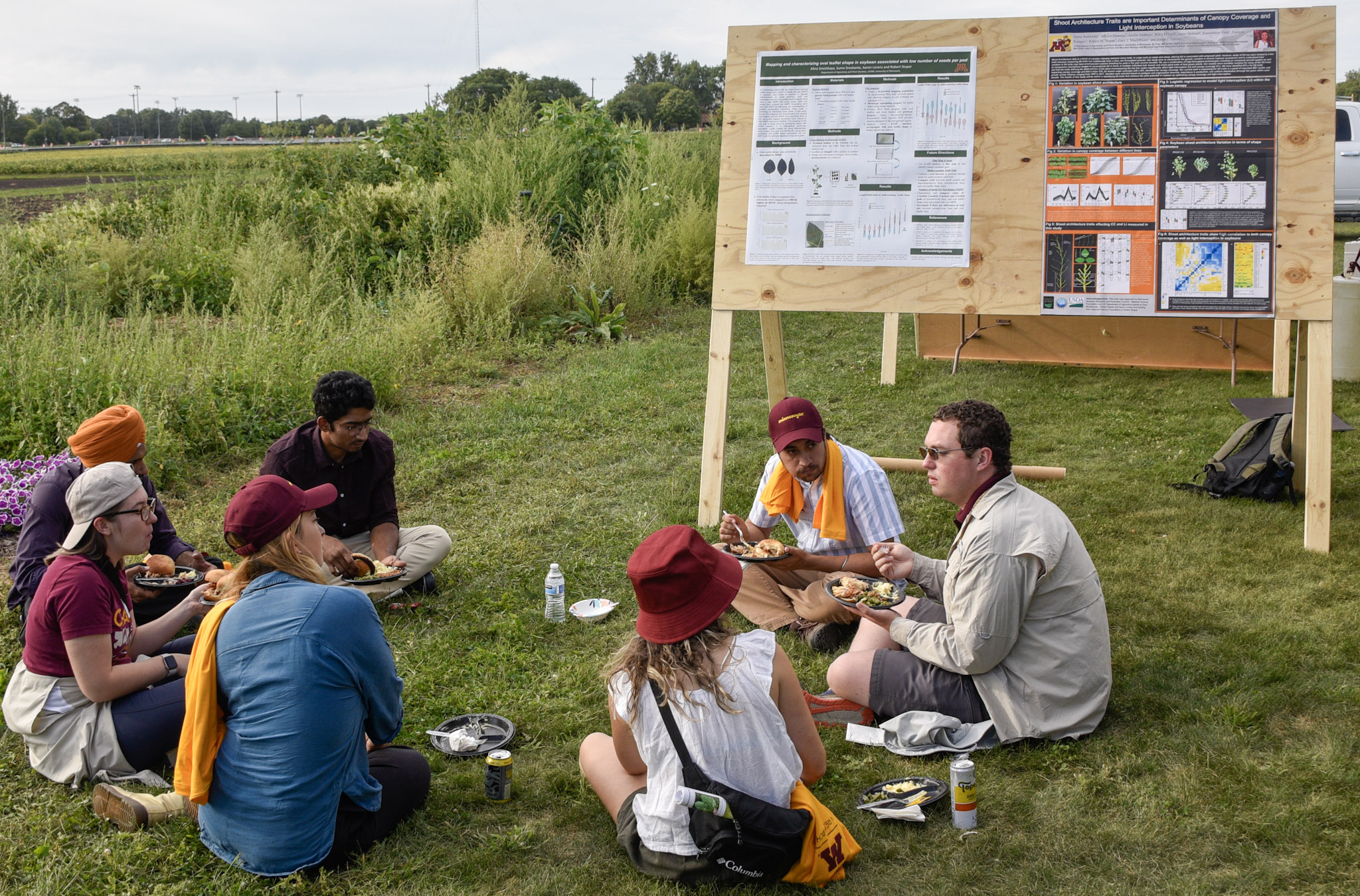 several people sit in grass eating in front of scientific posters