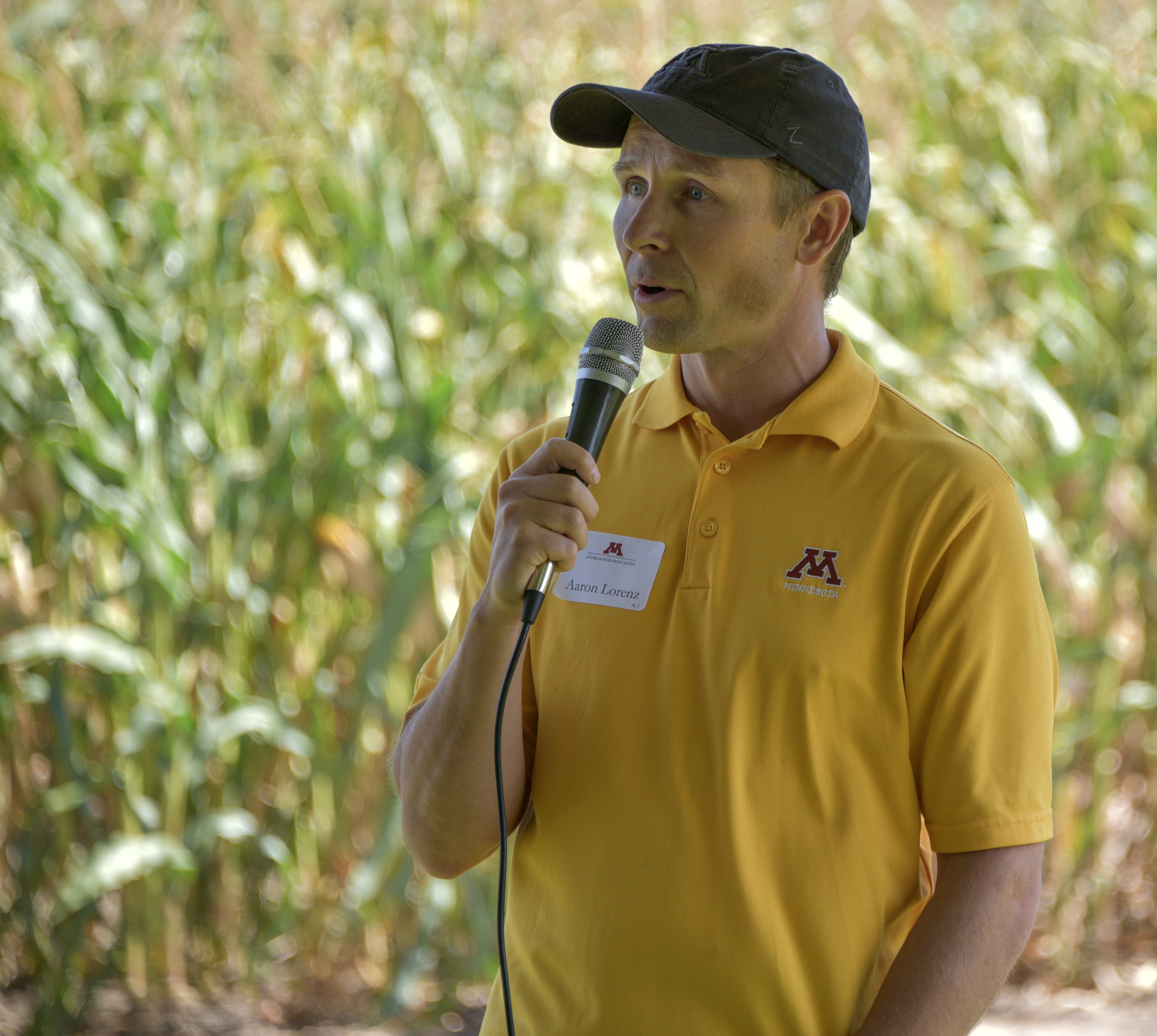 Aaron Lorenz, in a yellow shirt and baseball cap, speaks to the attendees in front of a field. 