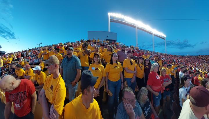 many people in gold Soybean Research Center tshirts stand outside watching the football game