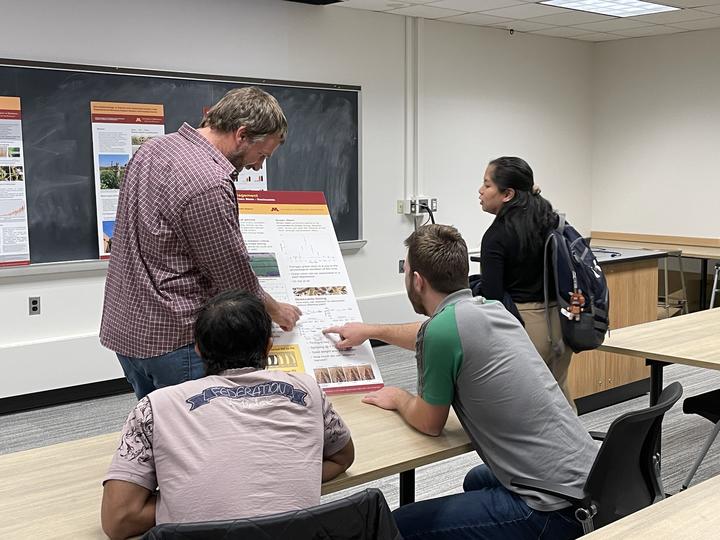 Man explains poster of his research to 3 students.